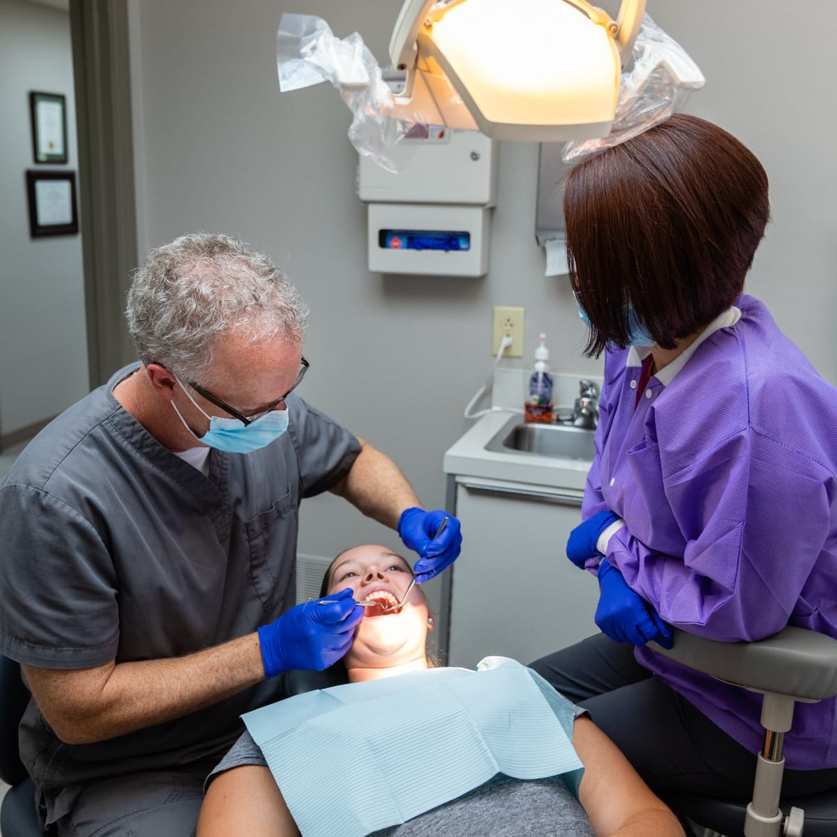 Dentist in gray shirt examining patient's teeth in clinical dental office setting.
