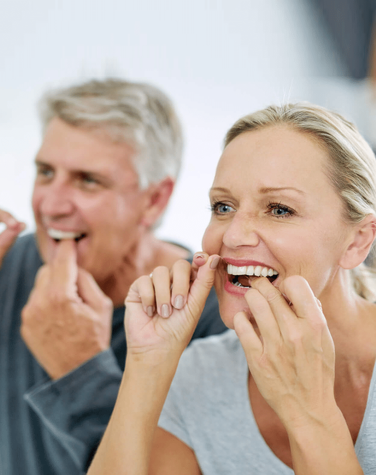 Man and woman flossing teeth together in bathroom, smiling at mirror.