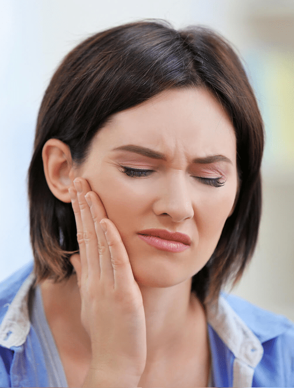 Woman in blue shirt touching her cheek, eyes closed, appearing to experience jaw or tooth pain indoors.